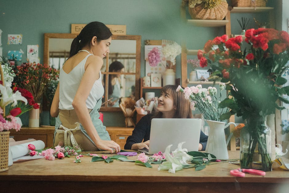 A female florist and a customer in a flower shop with green walls, engaging in a conversation about floral arrangements. The florist, wearing a beige apron over a white sleeveless top, is leaning acro