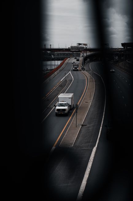 A view of a multi-lane road with a white delivery truck and several cars traveling amidst overcast weather, seen through a narrow gap between dark vertical bars or slats, creating a framing effect. In