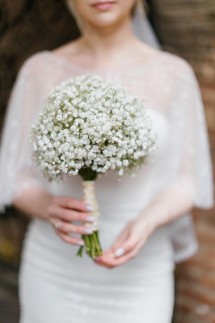 A bride holds a round bouquet of delicate white gypsophila flowers with small, ruffled petals, wrapped with a cream-colored ribbon around the stems. She is dressed in a lace wedding gown with sheer, e