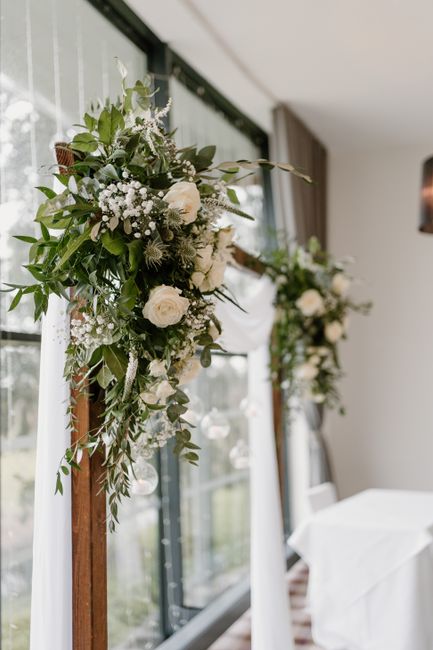 A floral arrangement featuring cascading greenery and white flowers, including roses and baby's breath, adorns a wooden stand at a wedding venue. The bouquet is lush, with a mix of glossy green leaves