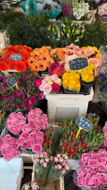 A vibrant display of freshly cut flowers at a florist stall, featuring bright orange gerbera daisies, soft peach and pink roses, and bundles of pink tulips arranged in white baskets and plastic sleeve